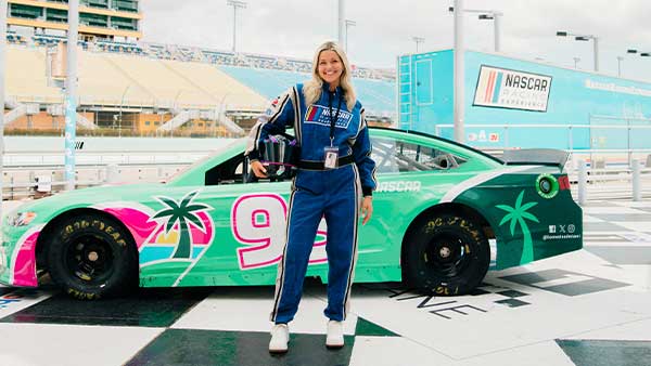 A woman wearing a blue NASCAR fire suit stands smiling on the checkered start-finish line beside a green and pink NASCAR stock car with palm tree graphics and the number 9, inside a race track facility.